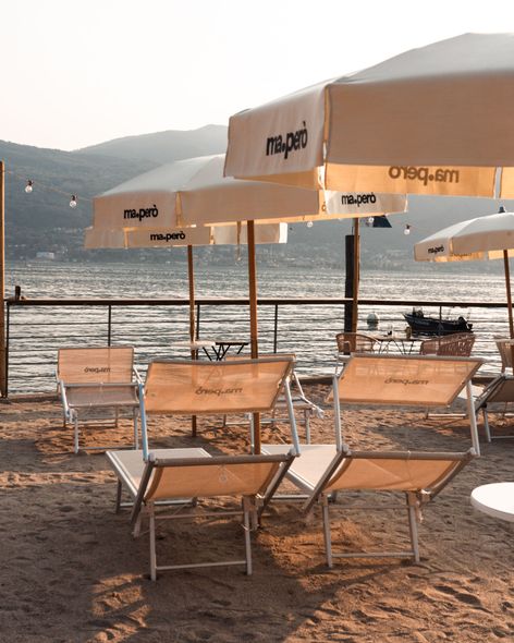 Scena di spiaggia con persone che prendono il sole e nuotano. Edifici e alberi costeggiano la riva sotto un tramonto colorato.