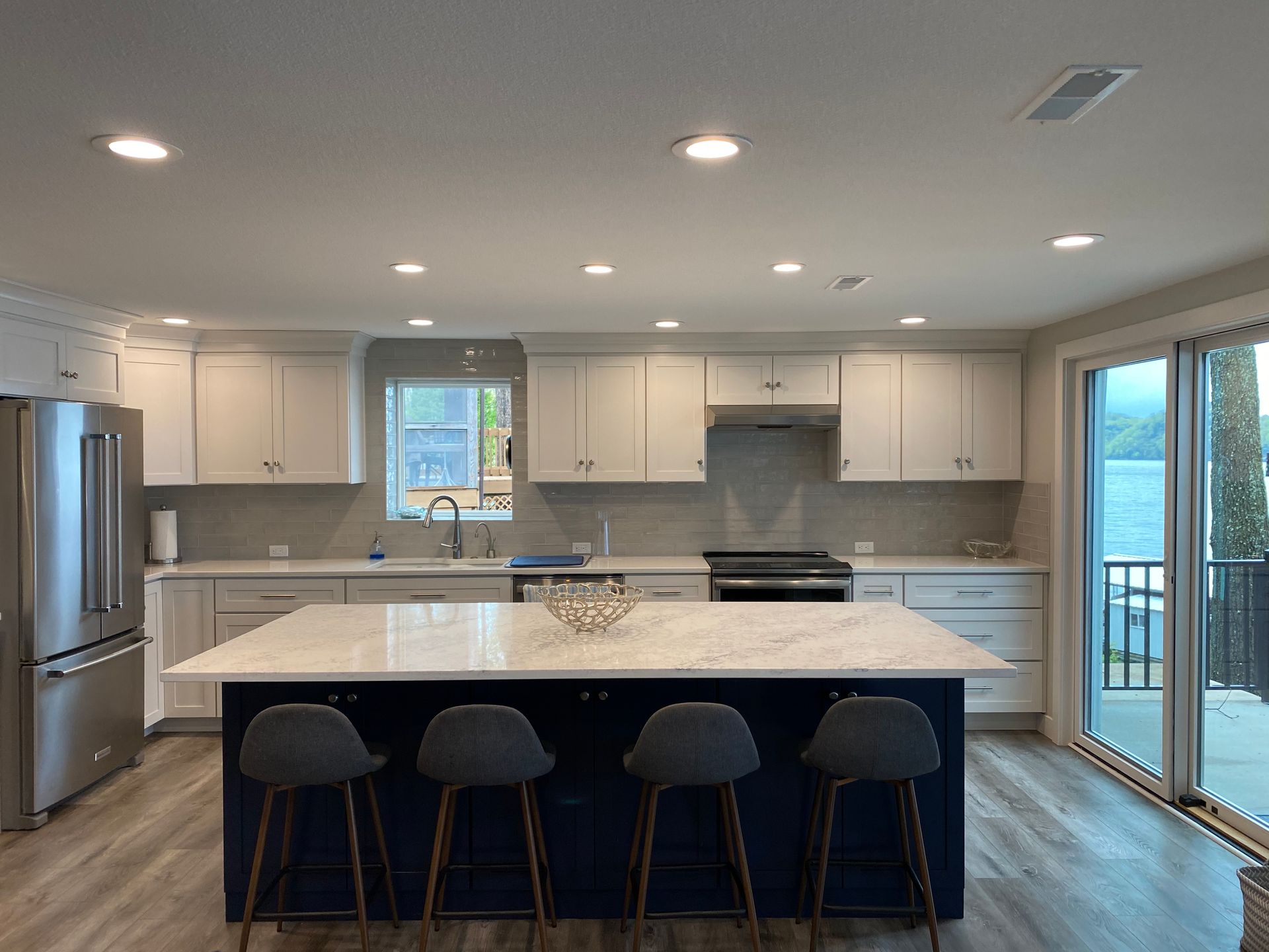 Bright modern kitchen with a large island, four stools, stainless fridge, and sliding glass doors.