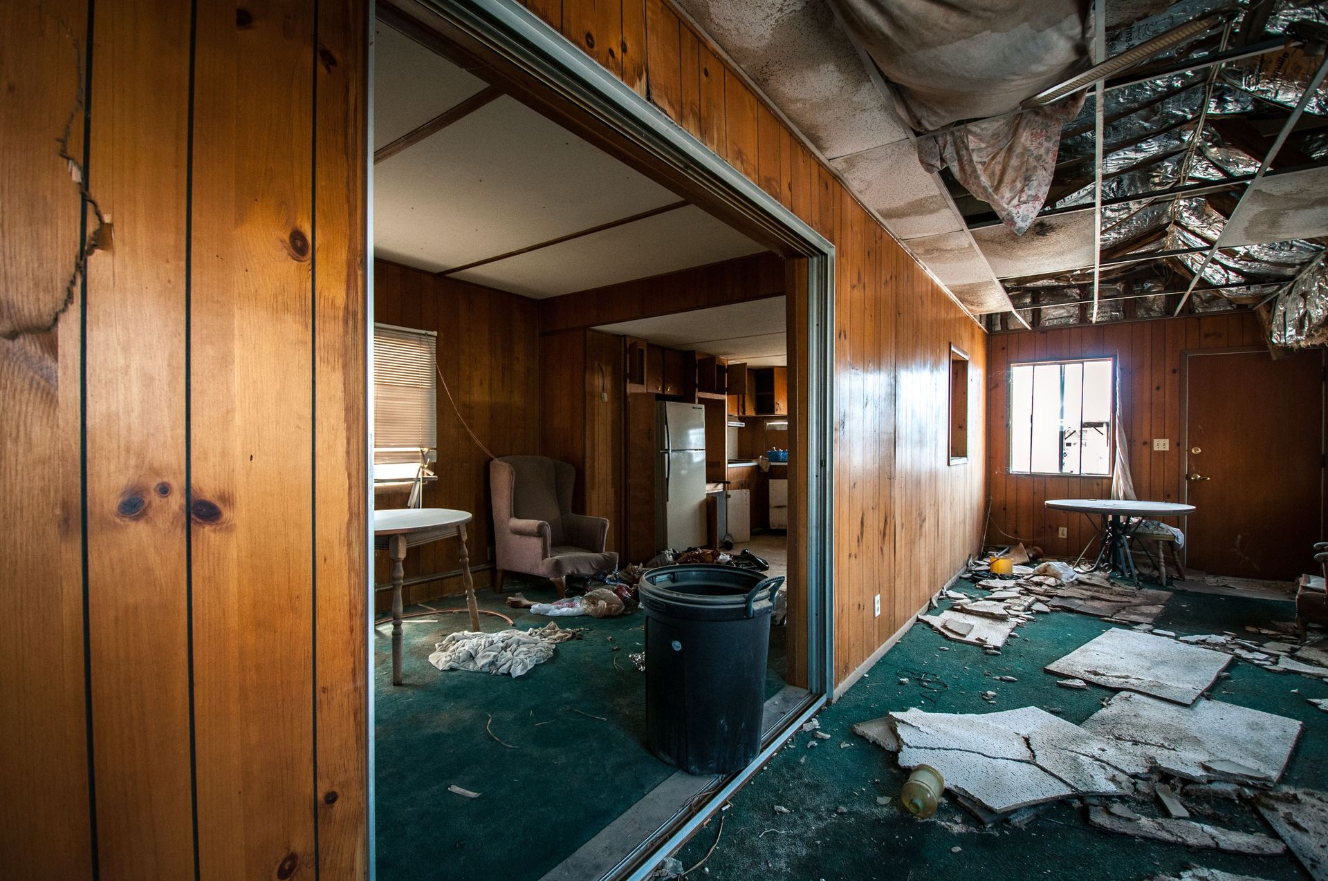 Abandoned, debris-strewn room with wood-paneled walls, torn carpet, and broken furniture