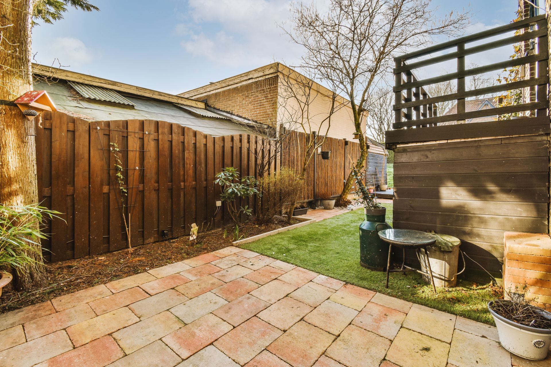 Backyard patio with wood fence, stone pavers, grass strip, and a raised deck on the right.