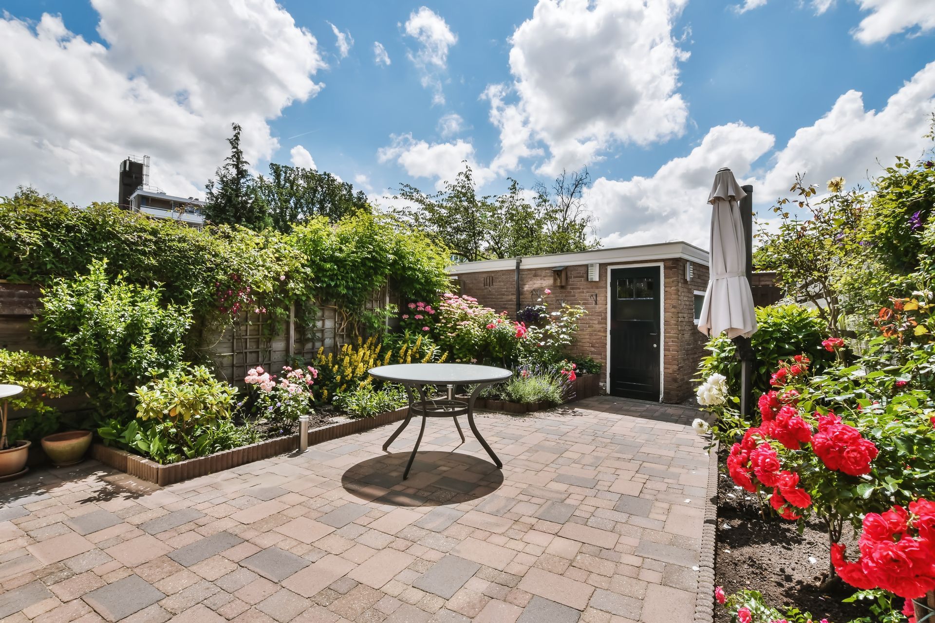 Patio garden with potted flowers, climbing greenery, round table, and closed umbrella under a bright sky