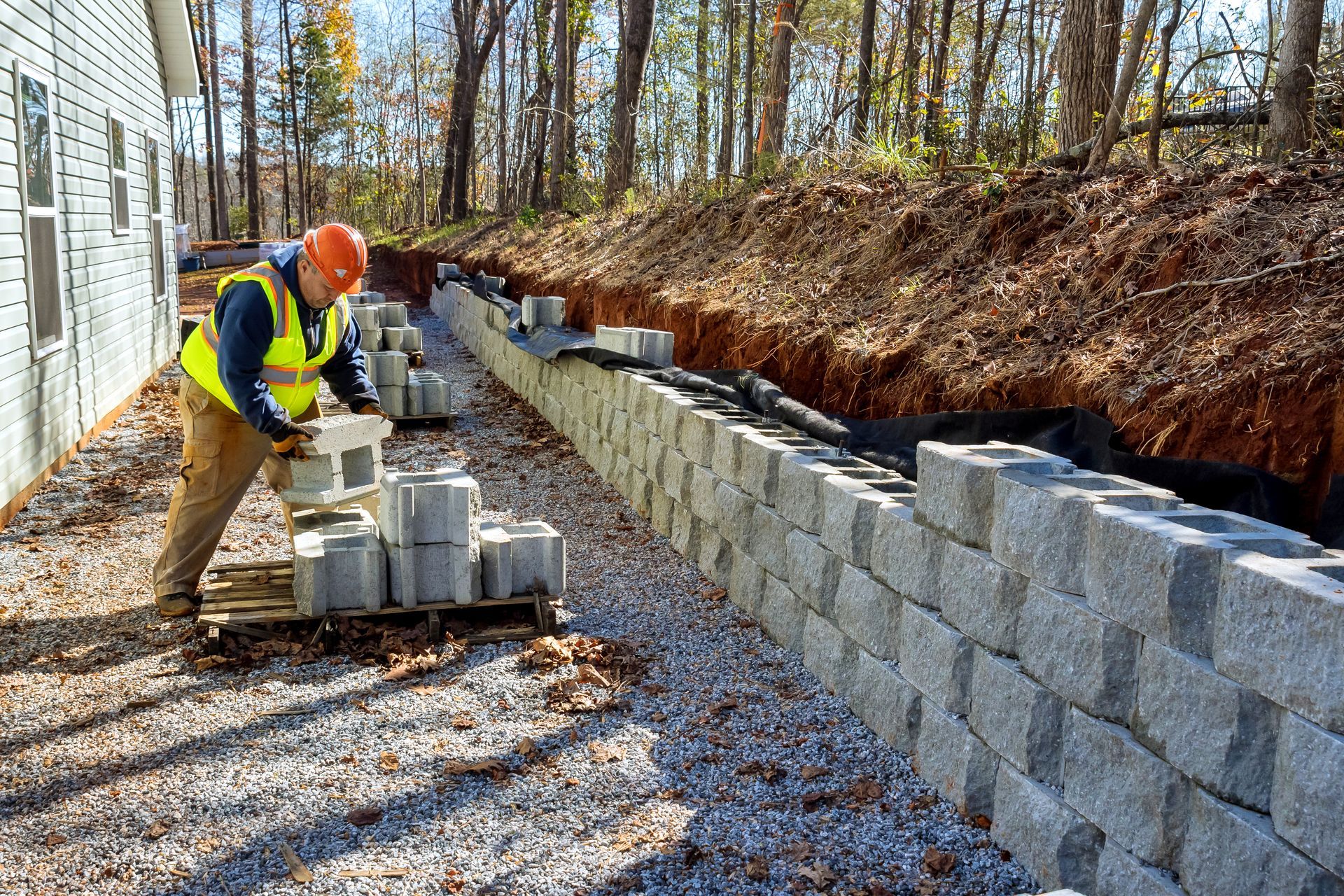 Construction worker stacking concrete blocks along a gravel path beside a retaining wall in a wooded area