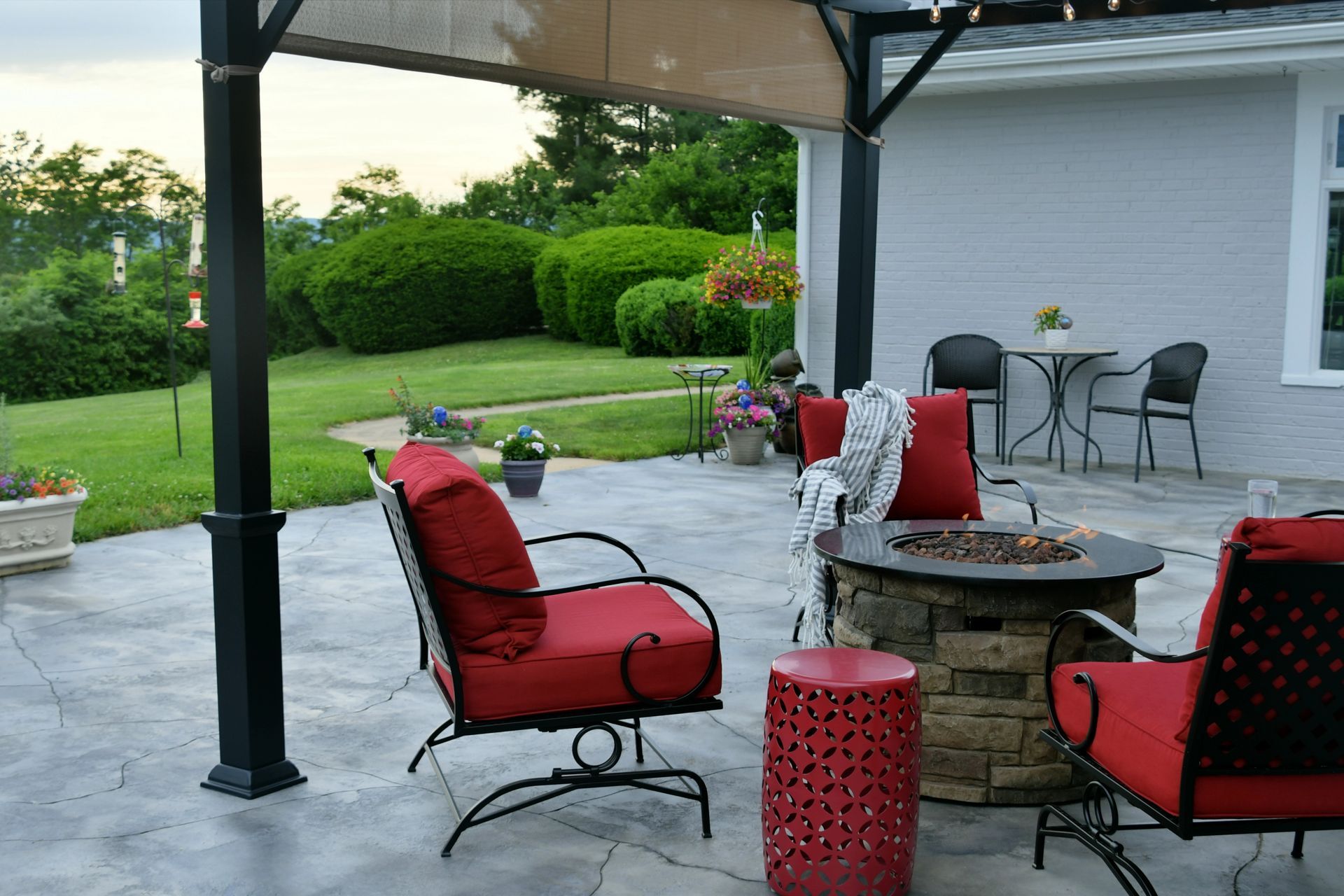 Covered patio with red cushioned chairs, stone fire pit, and green garden view.