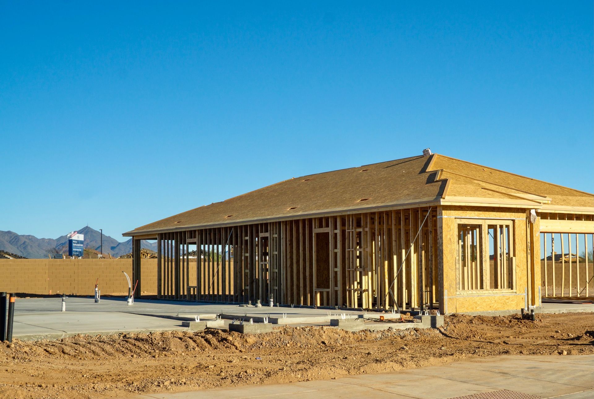 New wooden house under construction with exposed framing on a dirt lot under a clear blue sky