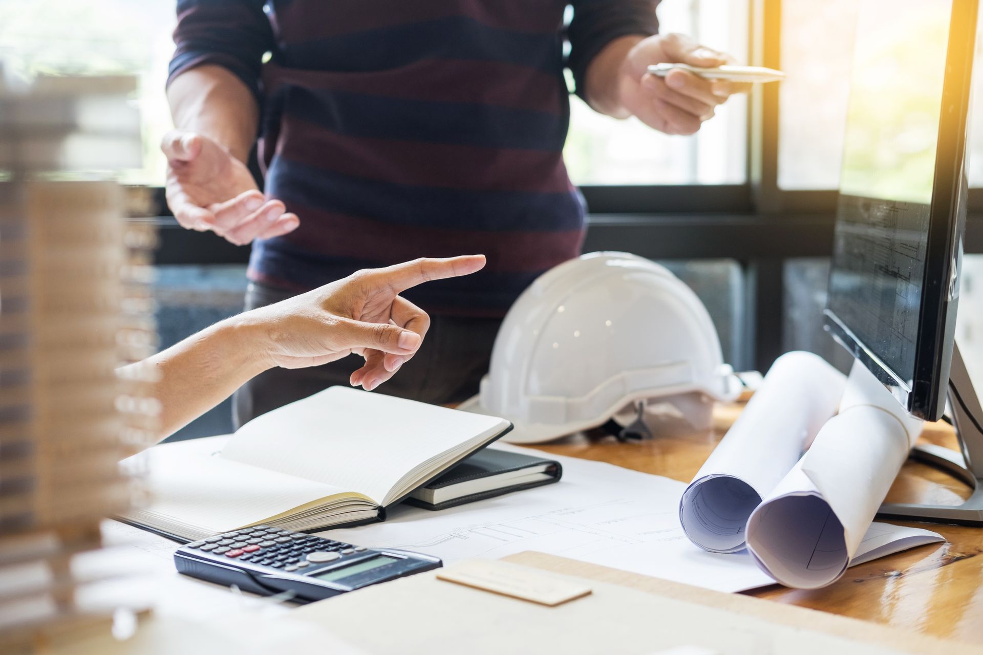 Two people reviewing plans at a desk with a hard hat, calculator, and rolled blueprints