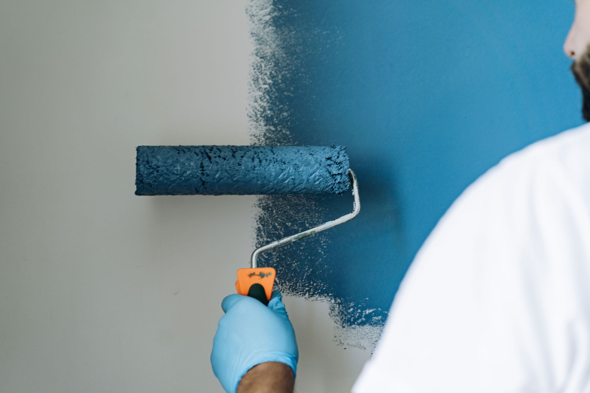 A person paints a wall blue with a roller, leaving part of the wall gray.