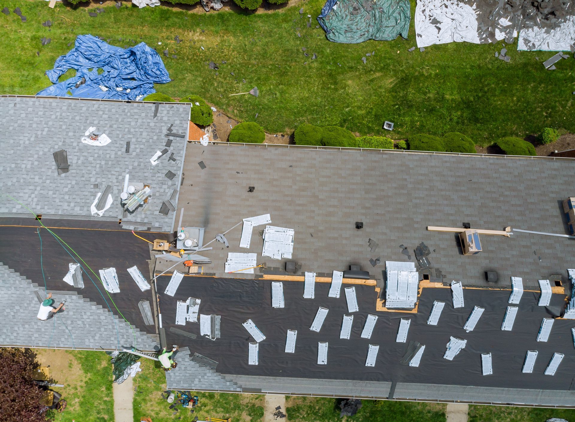 Aerial view of a roof under repair with white materials laid out and blue tarps on the grass nearby