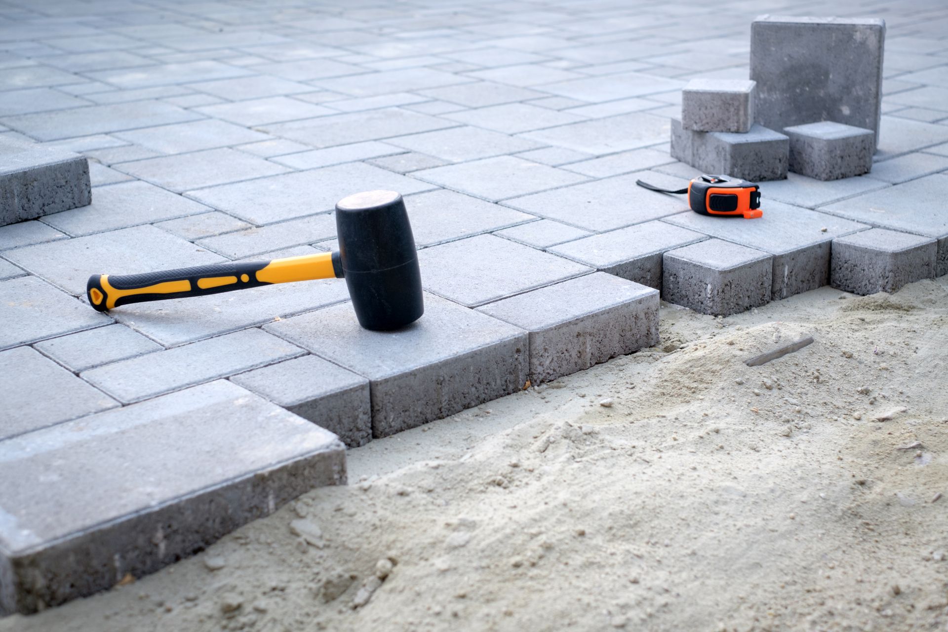 Paving project tools on sidewalk: black sledgehammer, orange tape measure, and concrete blocks.
