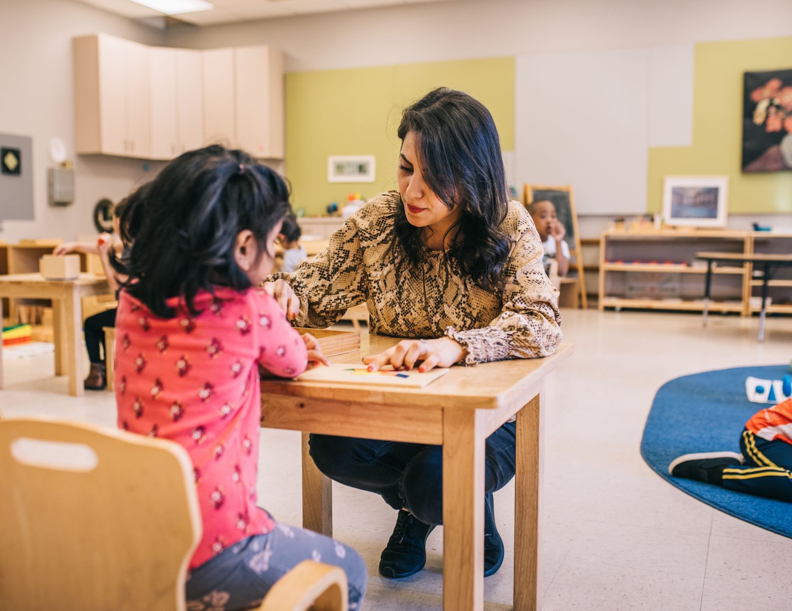 A woman is kneeling down next to a little girl at a table in a classroom.
