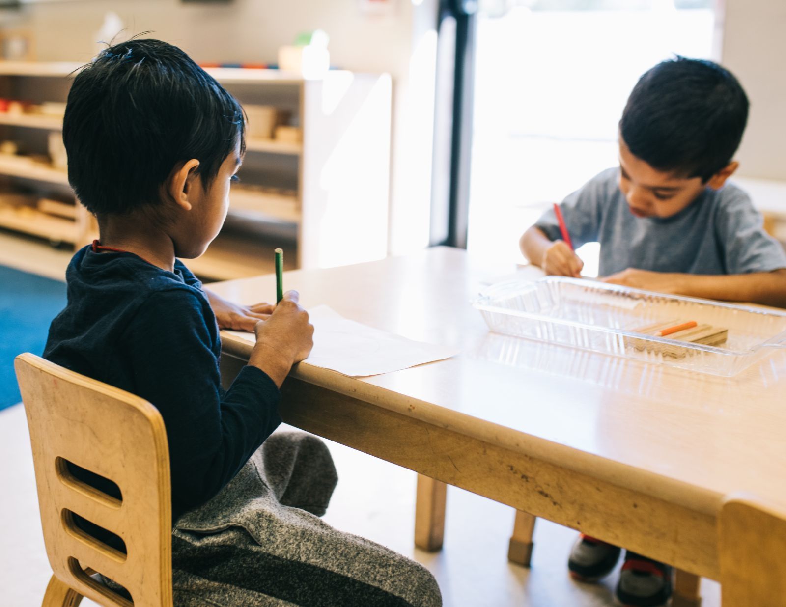 Two young boys are sitting at a table writing with pencils.
