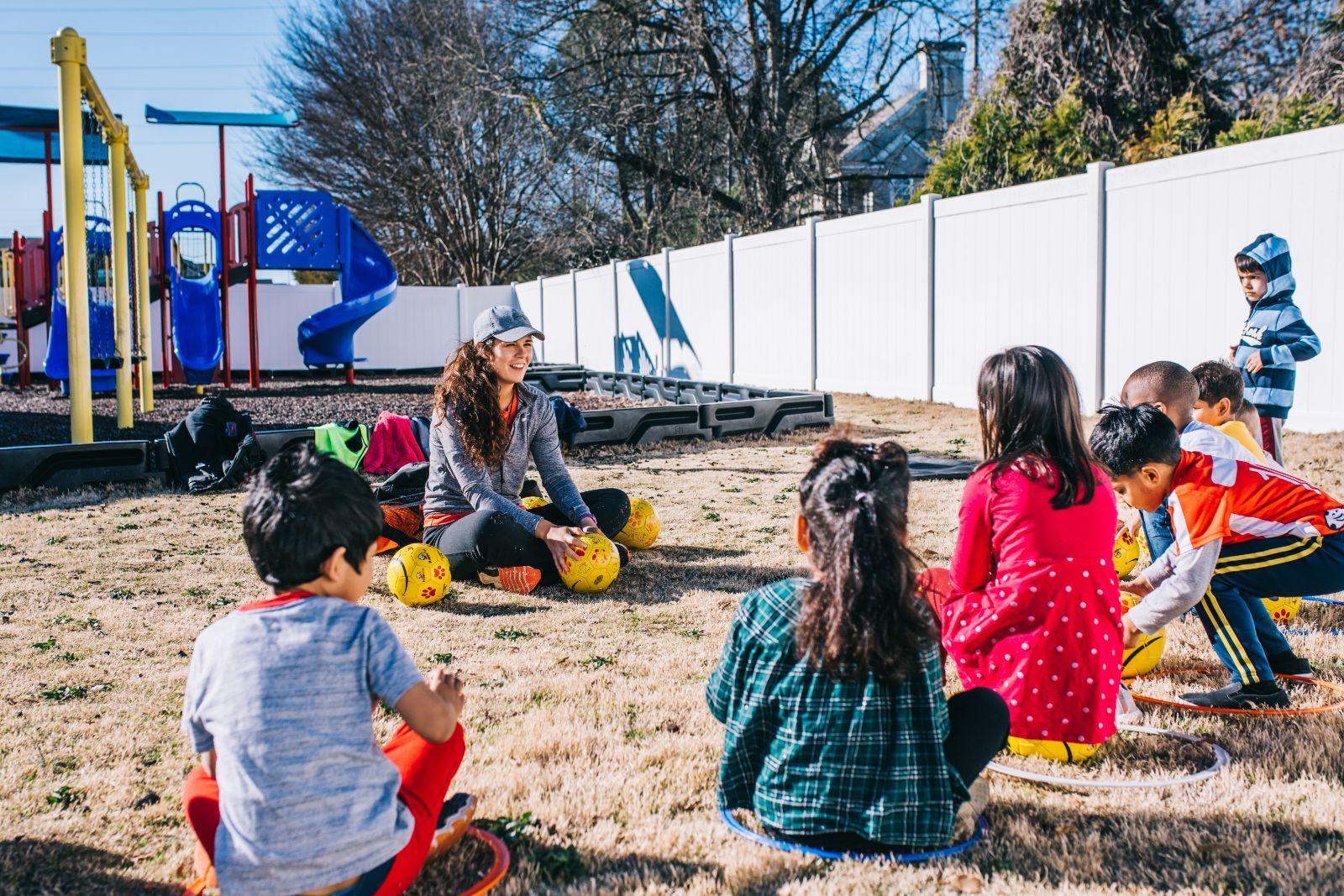 A group of children are sitting in a circle on the ground in front of a playground.