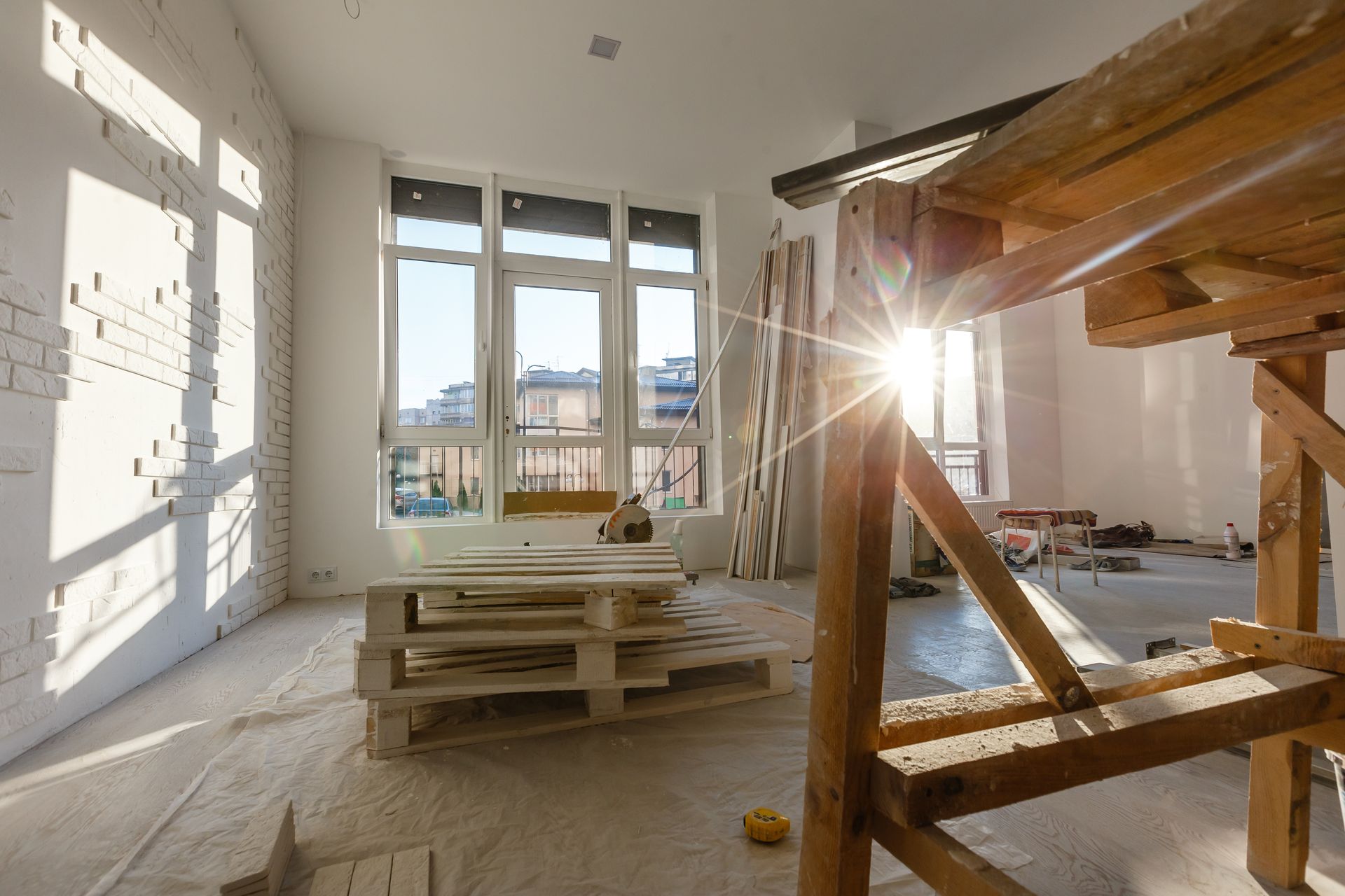 Sunlight streams through large windows into an unfinished room with white brick walls, wooden scaffolding, and pallets. Sunlight streams through large windows into an unfinished room with white brick walls, wooden scaffolding, and pallets.