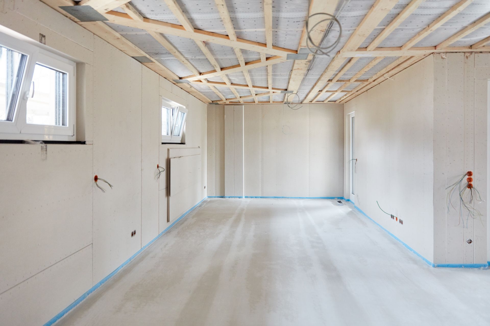 An interior room under construction with white plasterboard walls, a concrete floor, and exposed wooden ceiling beams. An interior room under construction with white plasterboard walls, a concrete floor, and exposed wooden ceiling beams.