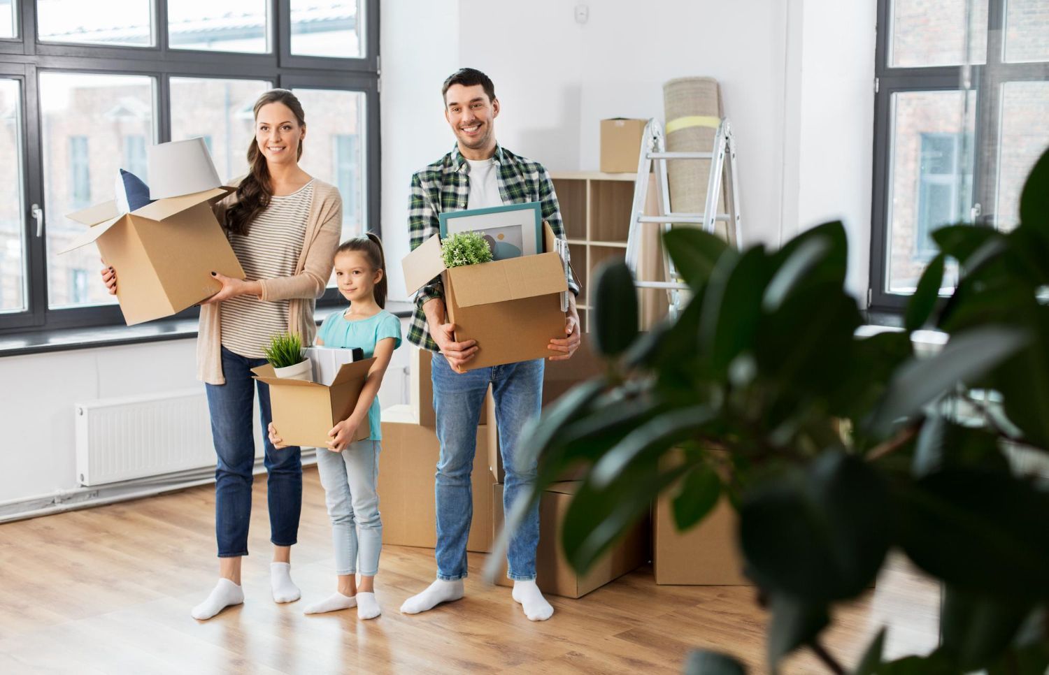 A family holds cardboard moving boxes and smiles in a bright, unfurnished room with large windows and wooden floors.