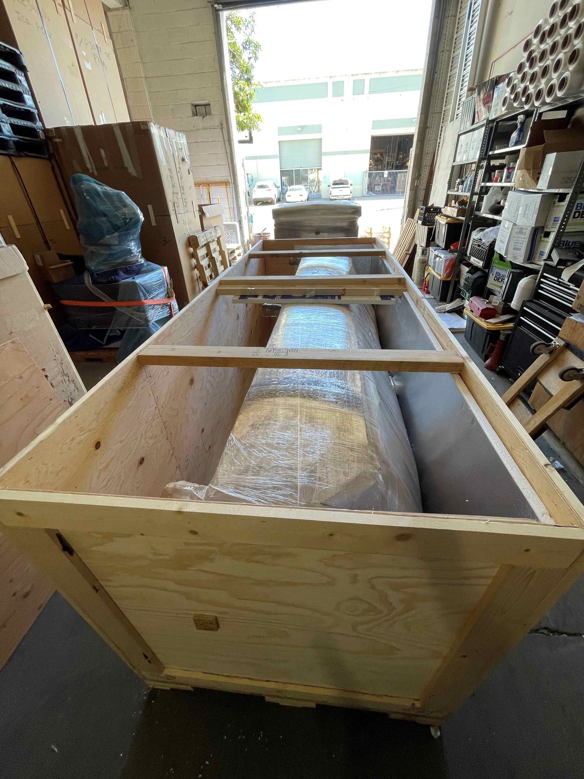 Large wooden shipping crate in a warehouse aisle, with foam-wrapped cylindrical equipment inside.