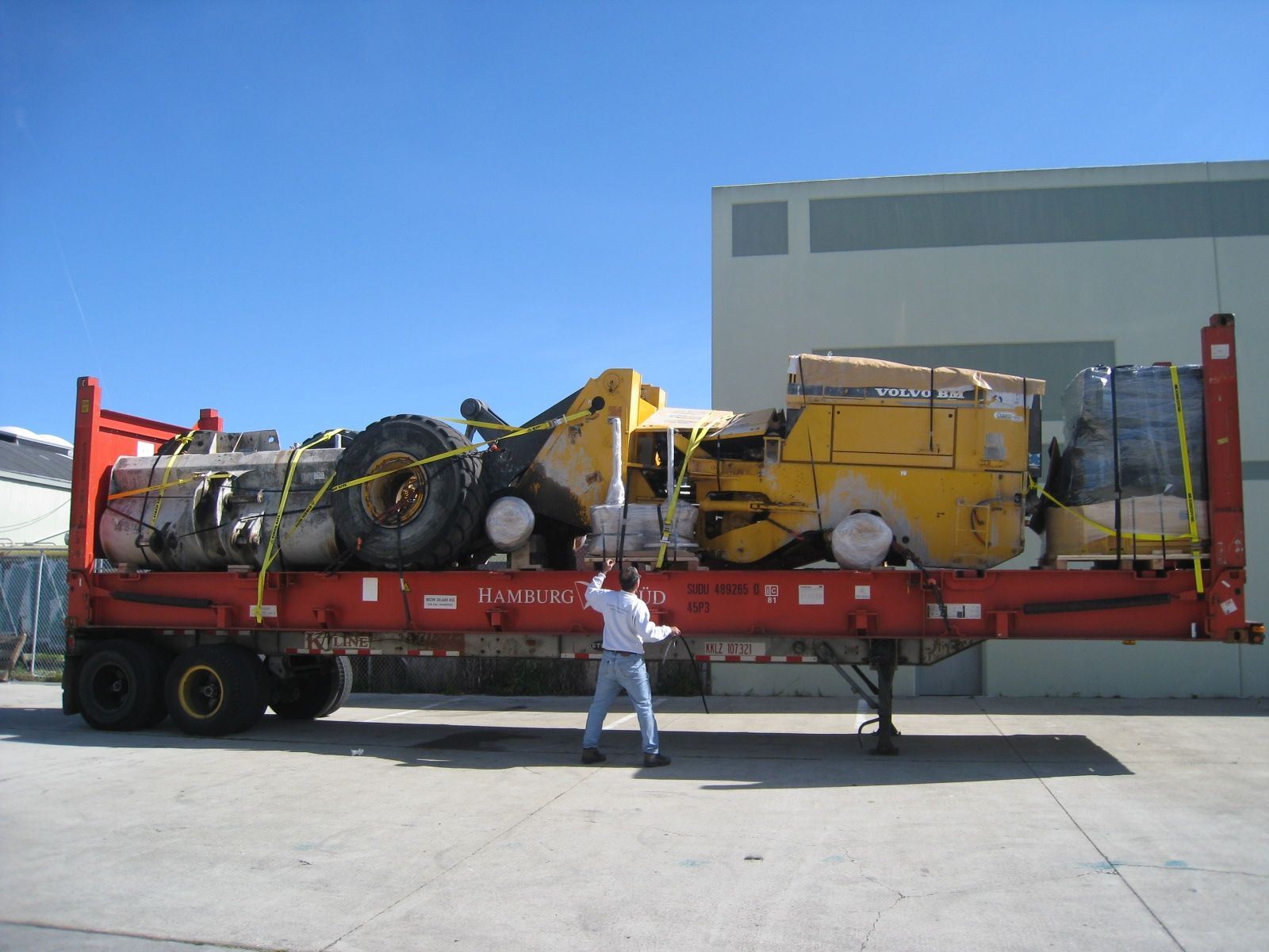 A person stands beside a flatbed trailer loaded with yellow construction machinery and industrial equipment outdoors.