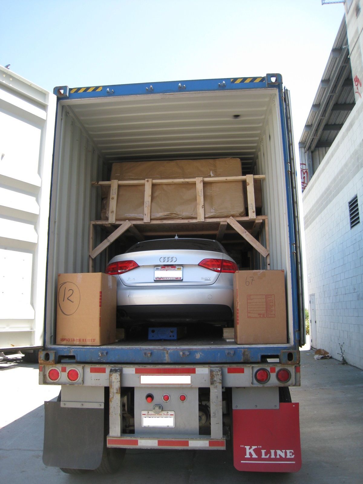 Silver car tightly packed in a shipping container, with wooden crates behind it at a warehouse dock.