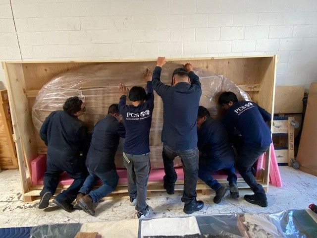 Six movers in matching dark blue uniforms work together to carefully slide a large, wrapped object into a wooden crate.