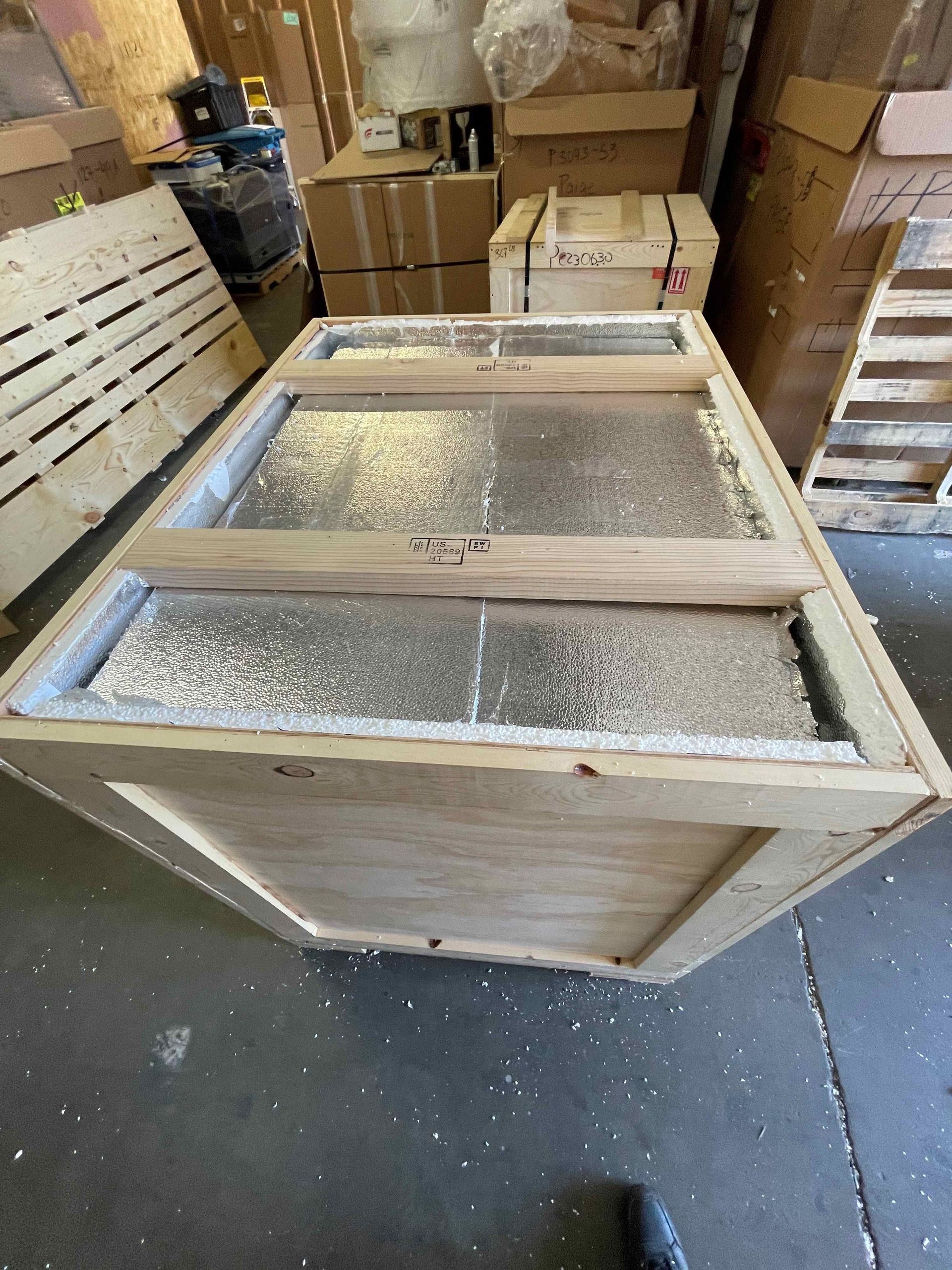 Wooden crate with a frosted glass top, sitting in a warehouse among stacked boxes.
