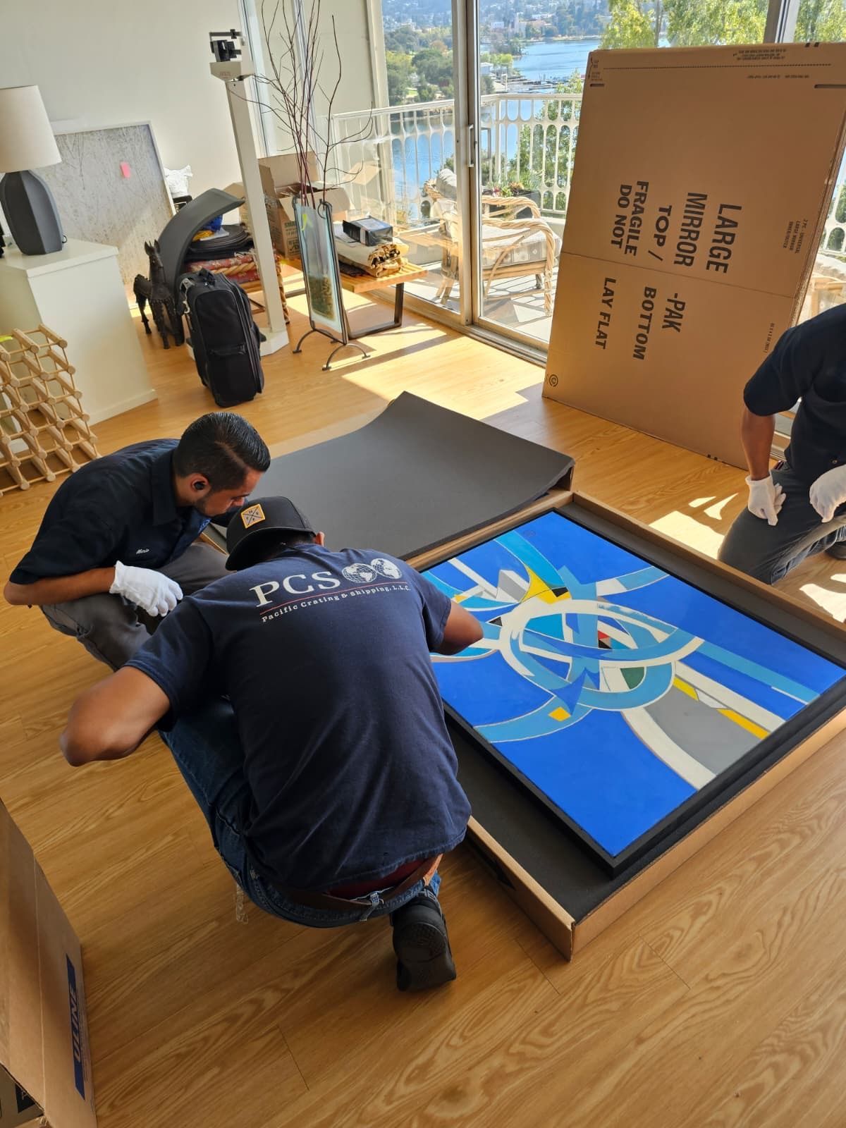 Two workers kneel over a framed colorful artwork in a bright room with a wood floor.