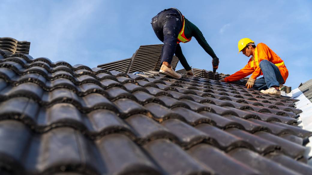 Two workers in safety gear repairing a dark tiled roof outdoors under a blue sky