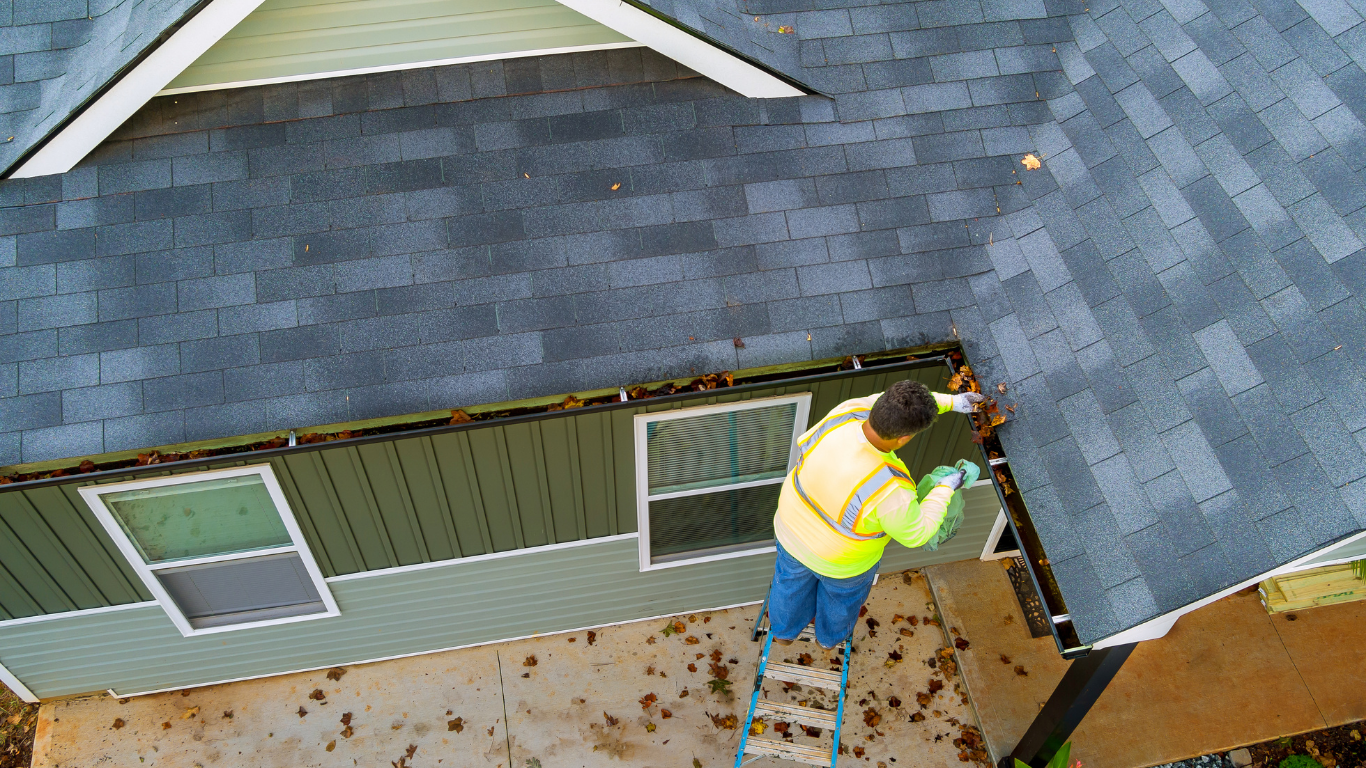 Worker in a yellow safety vest installing gutters on a gray-shingled roof.