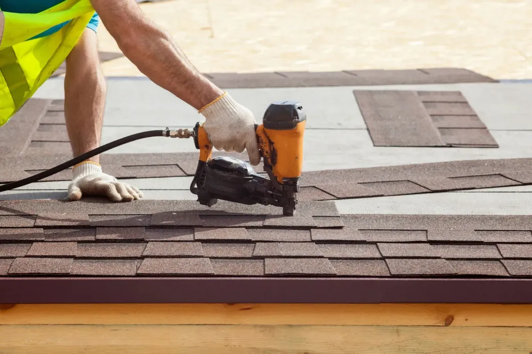 Worker using a nail gun to install roof shingles on a house roof