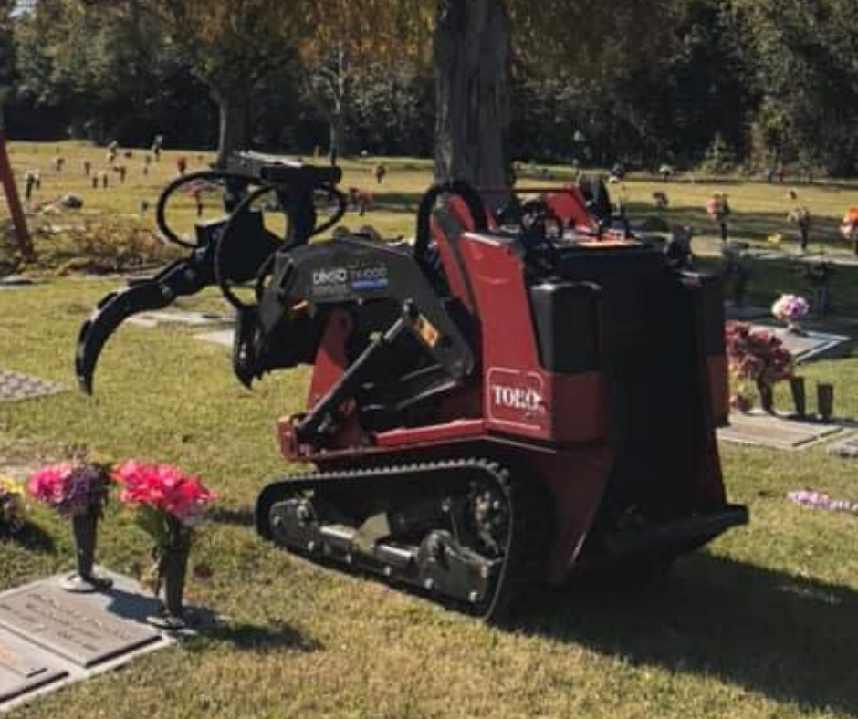 A red tractor is parked in a cemetery next to a tree.