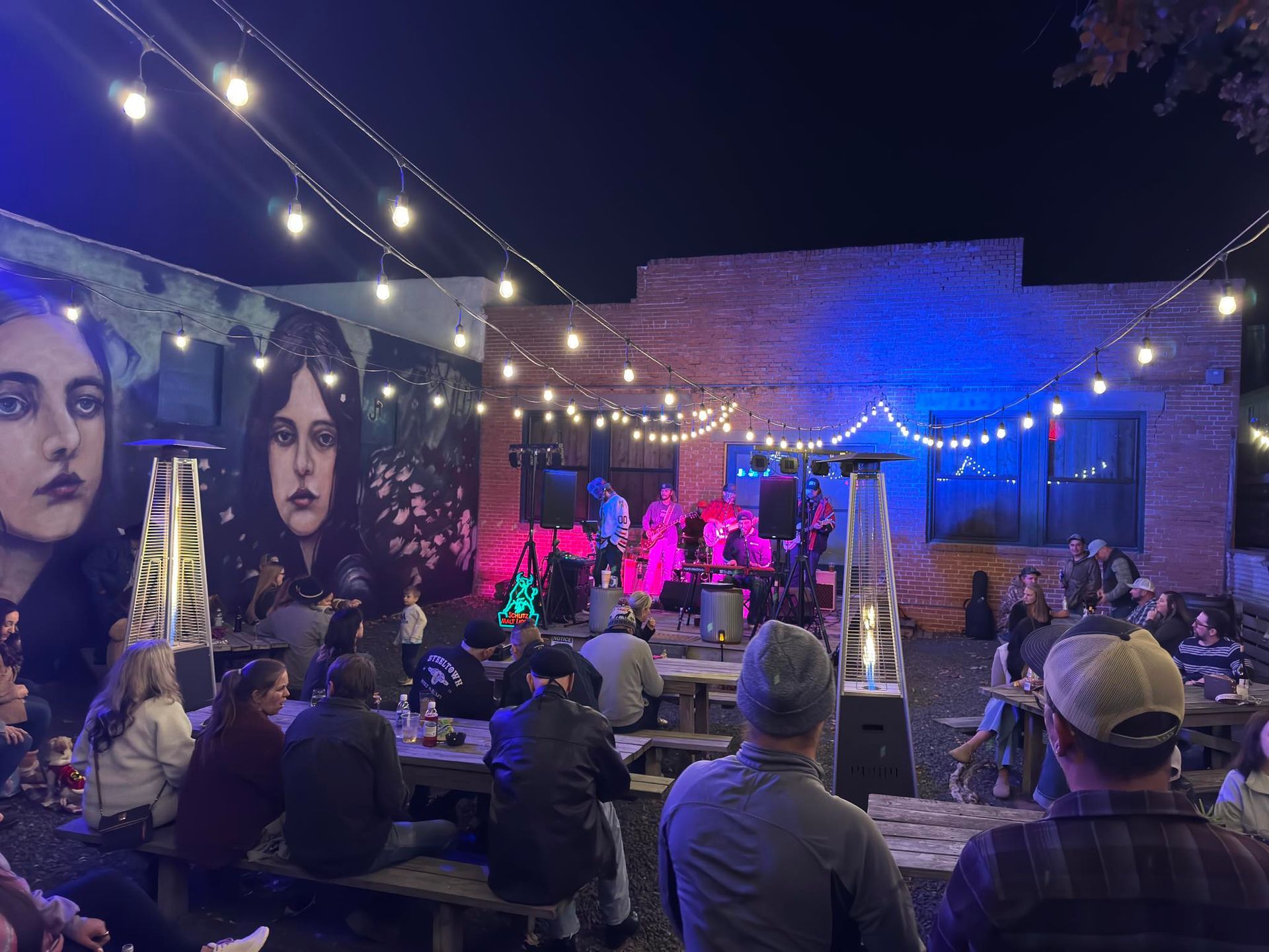 Outdoor concert at night: band on stage under string lights, crowd seated at picnic tables, mural on the wall.