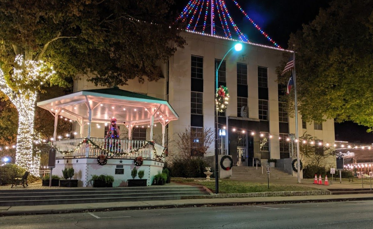 Night view of a gazebo and building decorated with Christmas lights in a town square.
