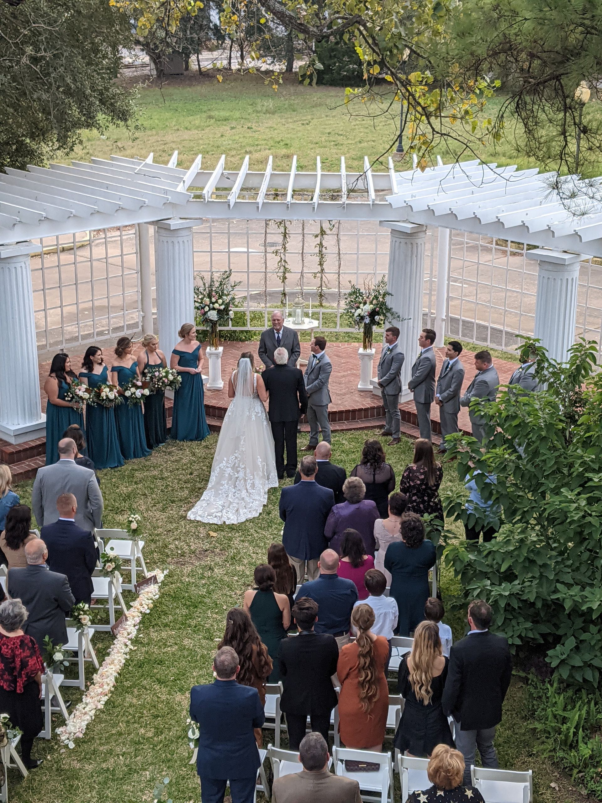 Wedding ceremony outdoors: Bride and groom at altar, flanked by bridesmaids and groomsmen. Guests in chairs face the couple. Green lawn, white trellis.