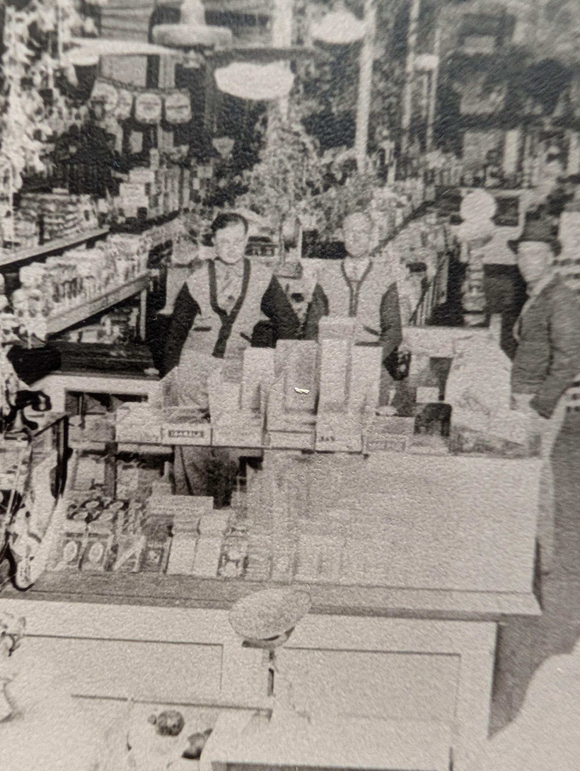 Two men behind a counter in a store, likely selling goods. Shelves filled with products. Black and white photograph.
