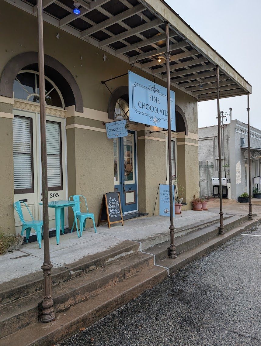 Exterior of a cafe with a teal sign, small table, and chairs. Building has a covered porch and arched windows.