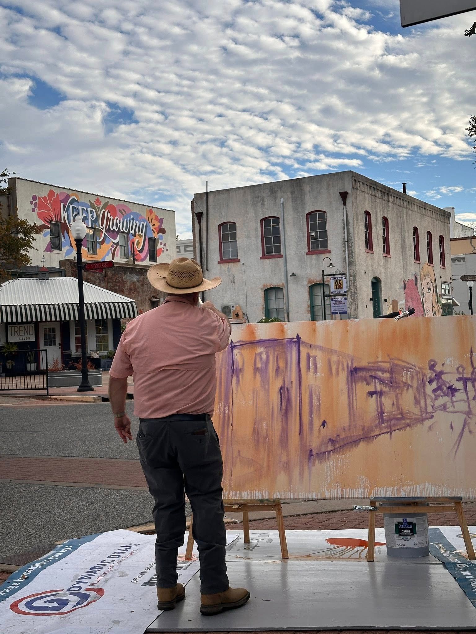 Artist painting outdoors; cityscape with buildings and blue sky; easel on a patterned ground.