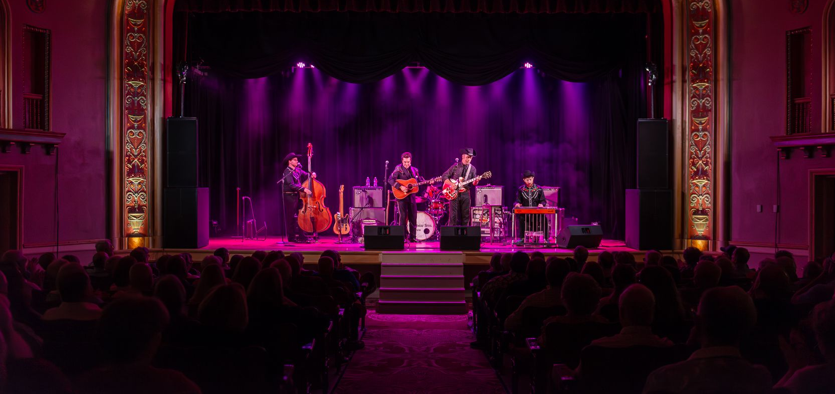 A band performs on a stage lit with purple lights in an ornate theater.