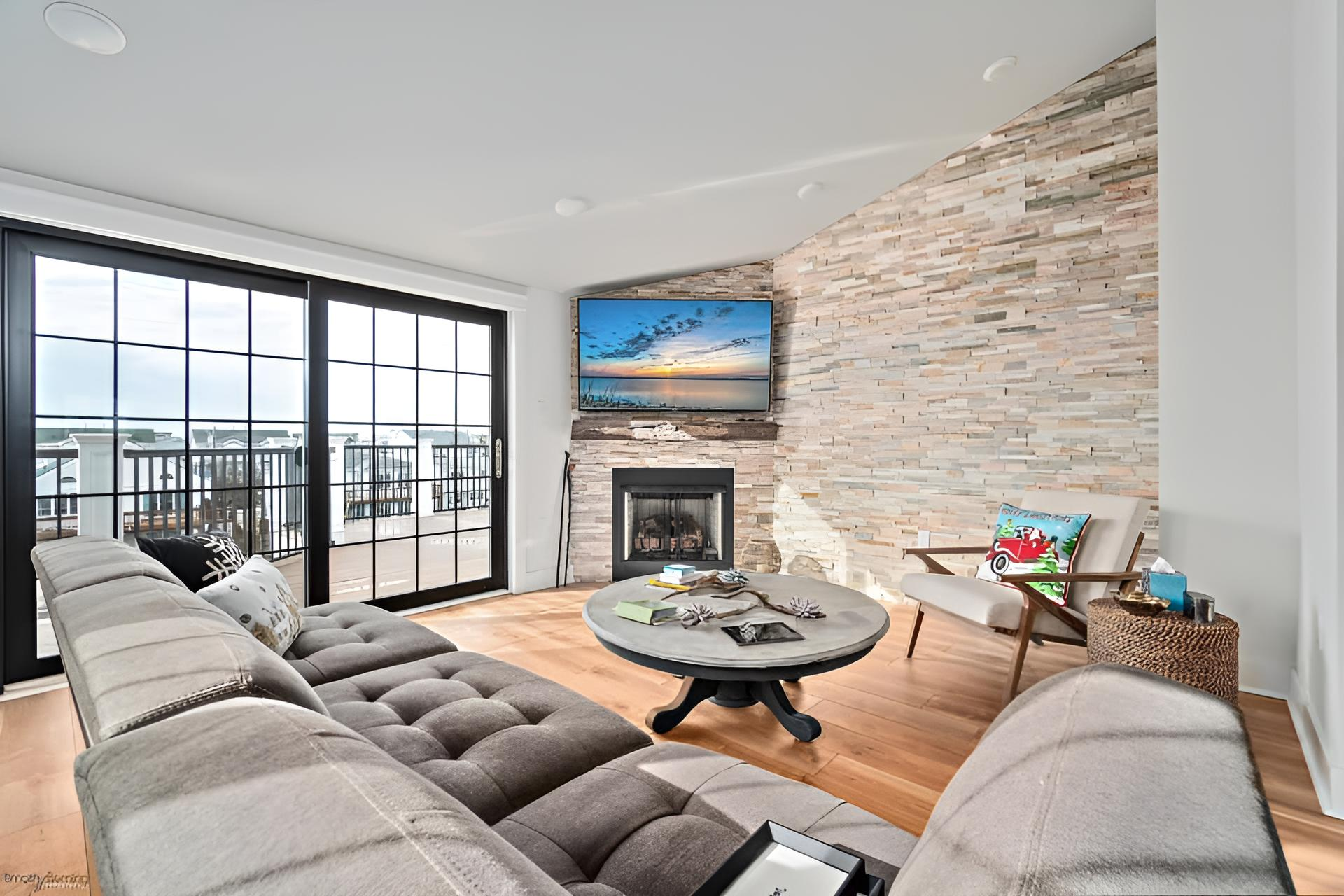 remodeled spacious living room with grey sectional, fireplace, TV, and large windows overlooking a deck by j.k. construction llc in avalon, new jersey