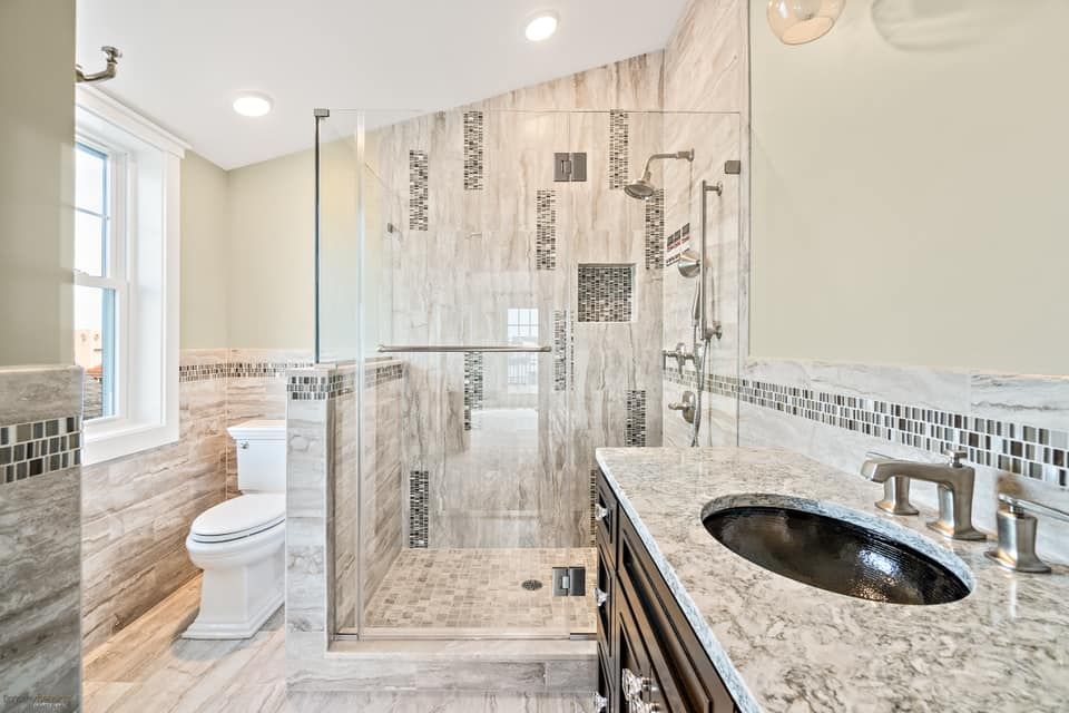 remodeled, modern bathroom with a glass shower, marble-like tile, dark vanity, and a toilet near a window in ocean city, new jersey done by j.k. construction llc 