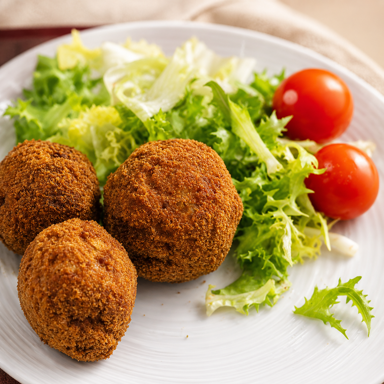 Tres croquetas empanizadas en un plato blanco con lechuga y tomates cherry.
