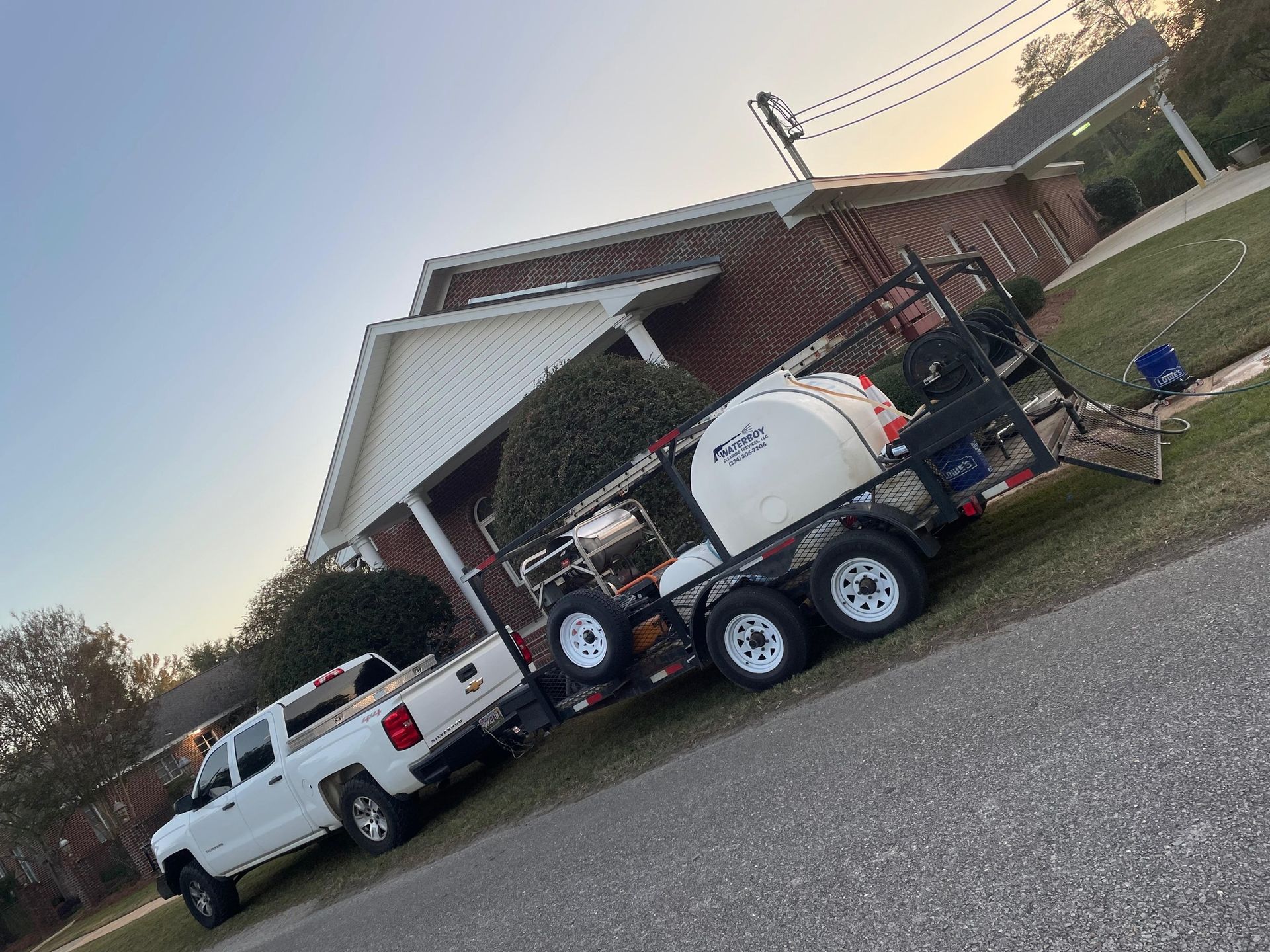 White pickup truck pulling a trailer with white tanks parked on a street near a brick house.