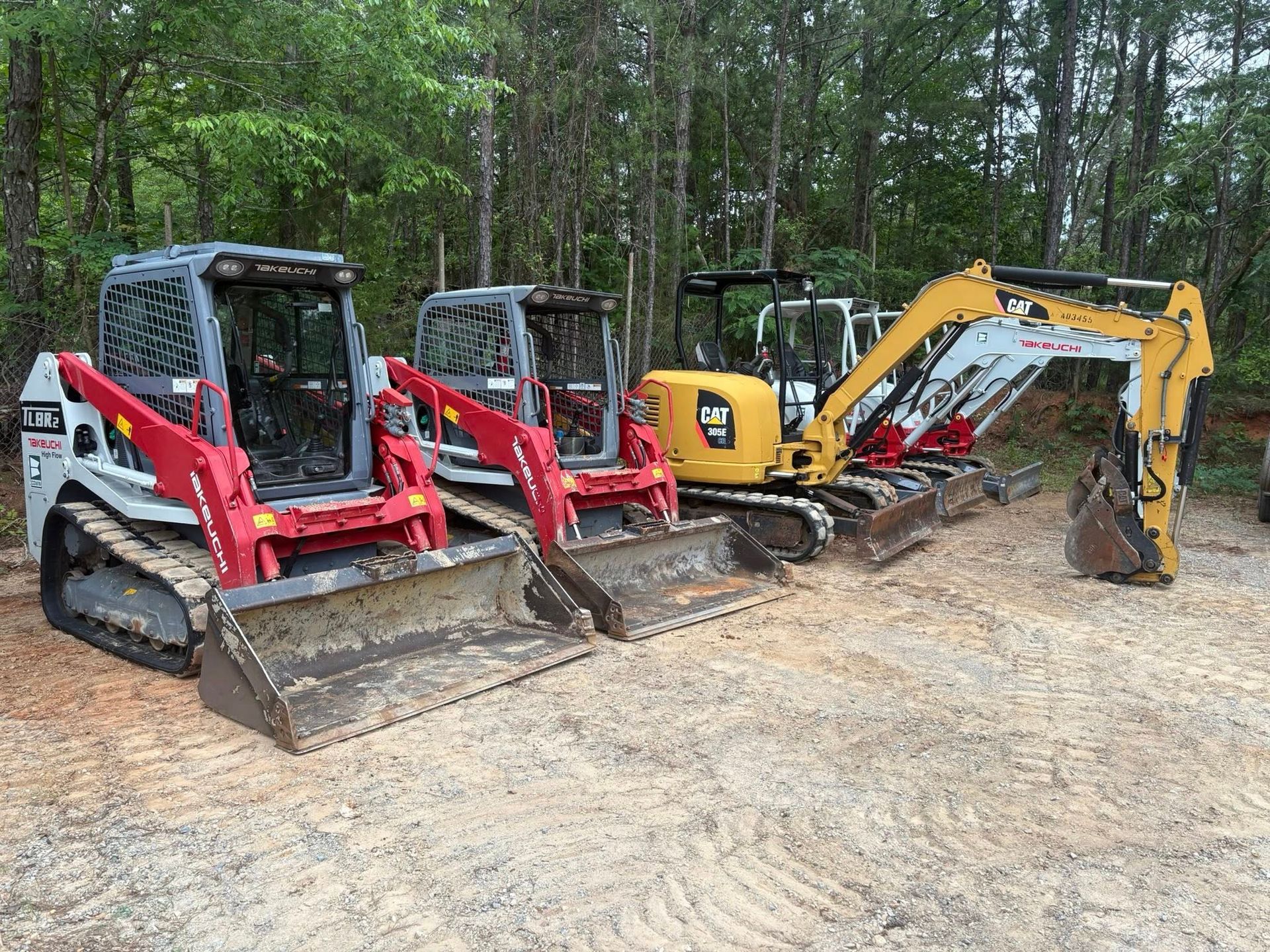 Three construction vehicles: two red and gray Track loaders and a yellow CAT excavator, parked in a wooded area.