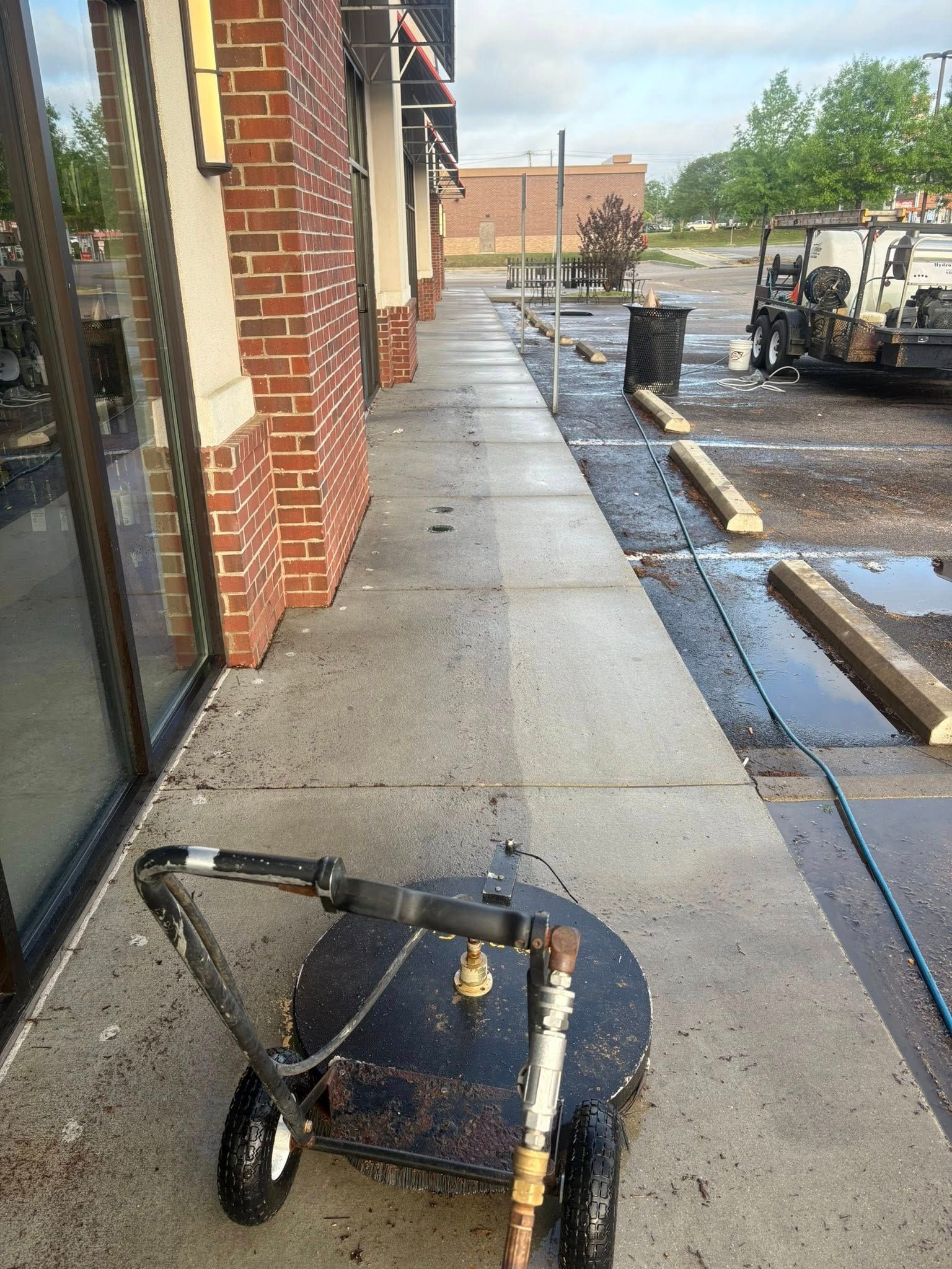 Sidewalk being pressure washed, showing before and after. Pressure washer in foreground, brick building at left.