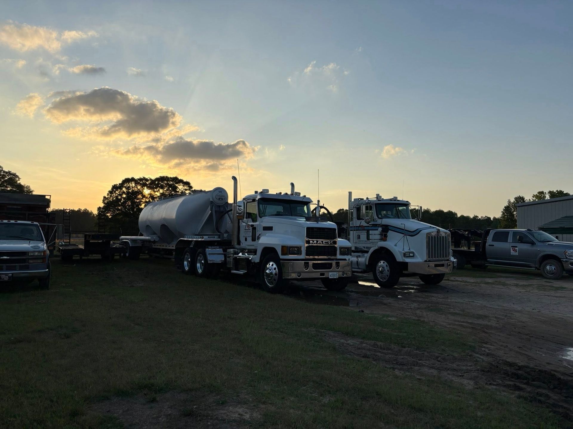 Two tanker trucks parked in a grassy lot at dusk, with a gray sky and few clouds.