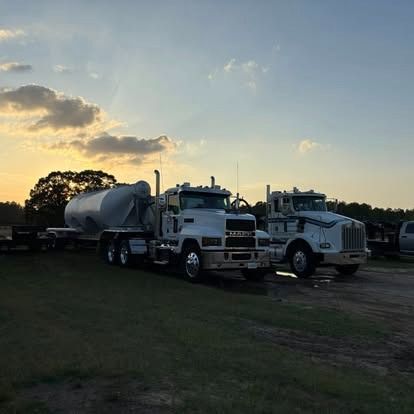 Two white semi-trucks parked on grass at sunset.