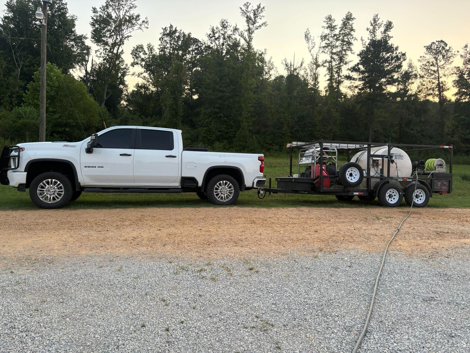 White pickup truck towing a trailer with two tanks and equipment on a gravel road, trees in the background.