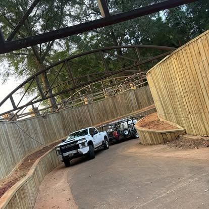 White pickup truck towing a trailer travels on a curved road, under a bridge and wooden wall.