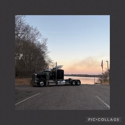 Black semi-truck parked near a body of water at dusk. Trees and sky in the background.