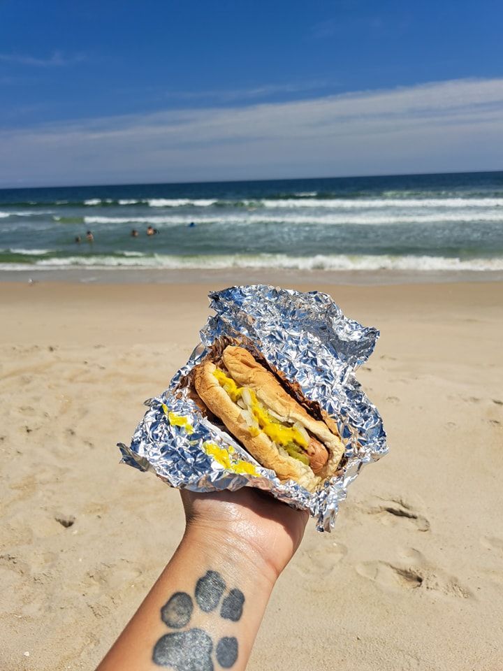 A person with a tattoo on their wrist is holding a hot dog on the beach.