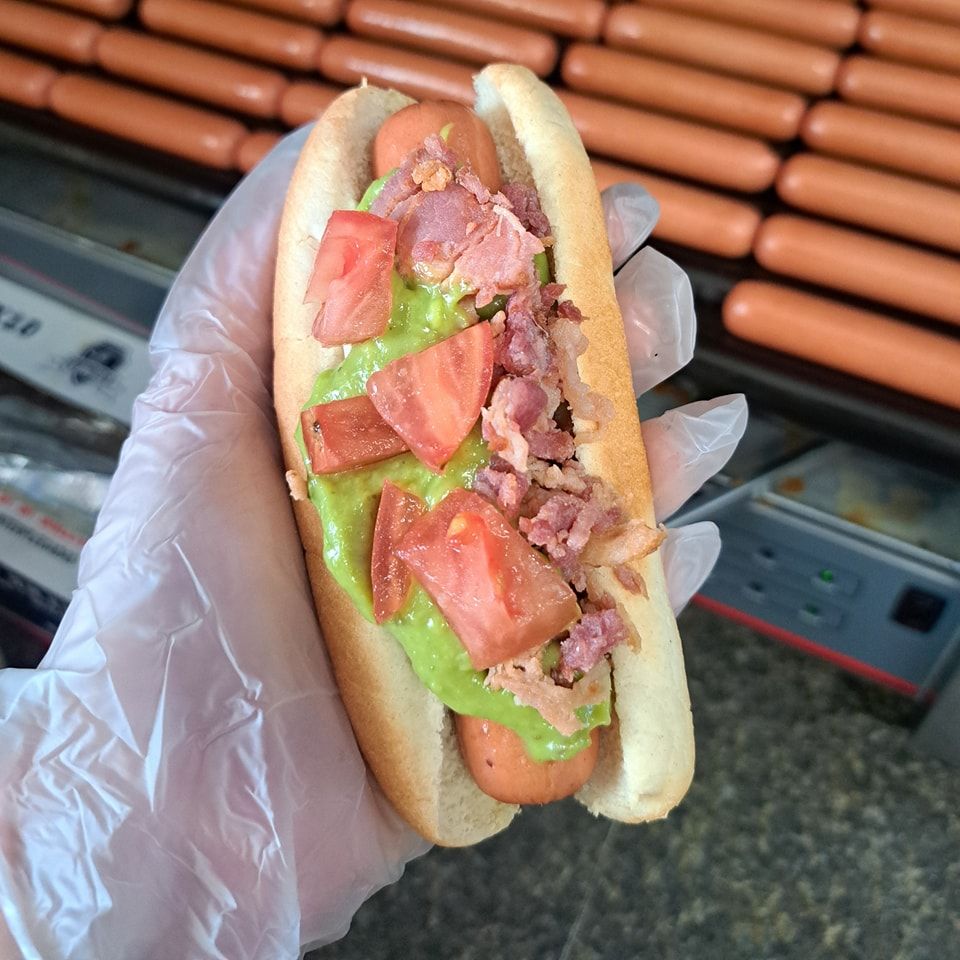 A person wearing gloves is holding a hot dog with tomatoes and bacon