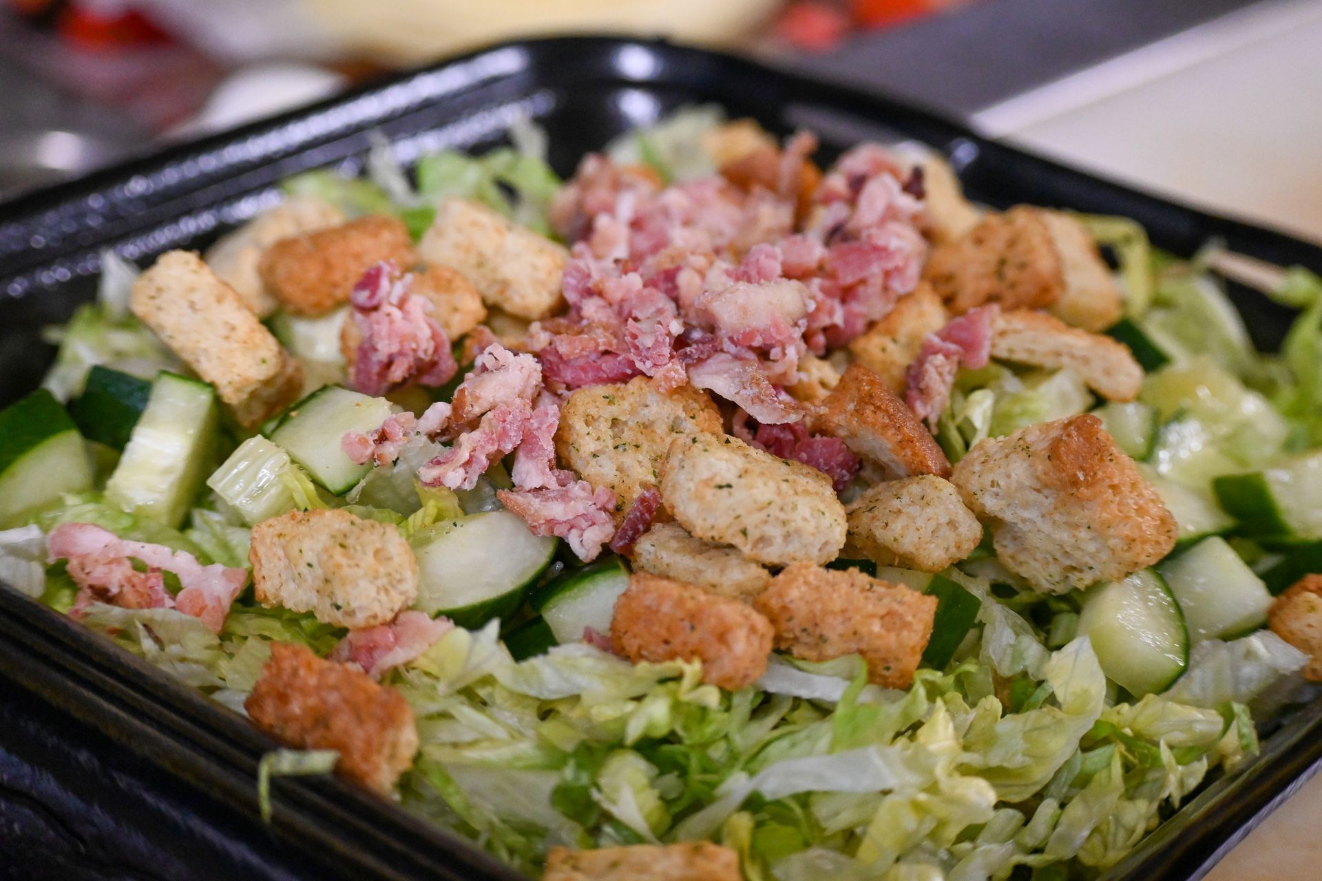 A salad with lettuce , croutons , bacon and cucumbers in a black container.