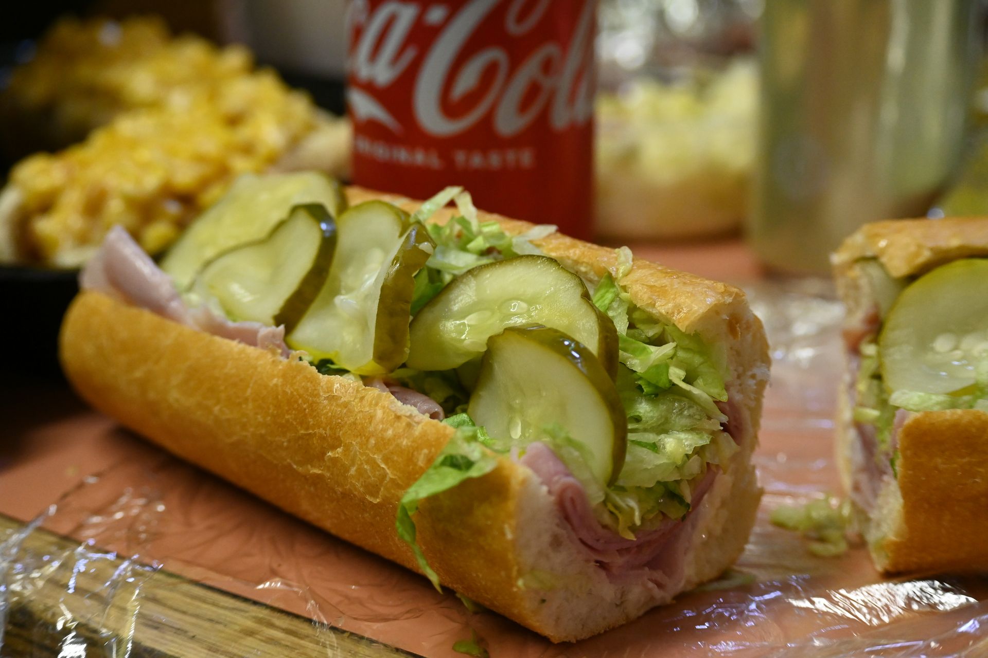 A sub sandwich with ham , lettuce , pickles and a coca cola can in the background.